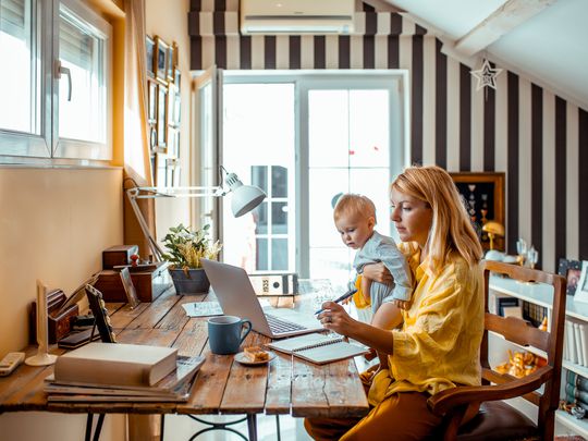 A woman sitting at a desk holding a baby and looking at a laptop