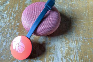 A red Chromecast propped against a pink, cloth-covered Google Home Mini.