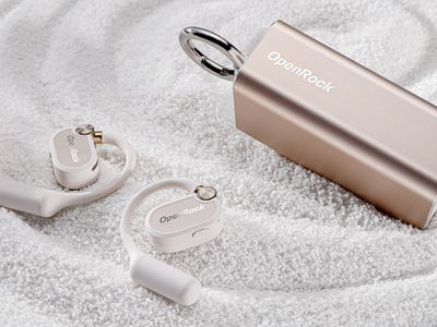 Pair of OpenRock X earbuds on white sand with charging case in metallic pink